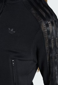Veste noire zippée avec le logo trèfle Adidas sur la poitrine et tissu en maille avec détail à trois bandes sur la manche.