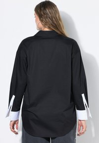 Donna con capelli lunghi che indossa una camicia nera oversize con polsini bianchi, in piedi di spalle contro un muro bianco.
