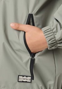 Mano nella tasca con cerniera di una giacca grigio chiaro con polsino elasticizzato, con un'etichetta nera "Original Denim Factory" vicino alla tasca.