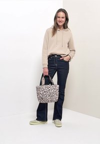 Femme portant un sweat à capuche beige et un jean foncé, tenant un sac fourre-tout à motif léopard, debout devant un fond blanc et beige clair.