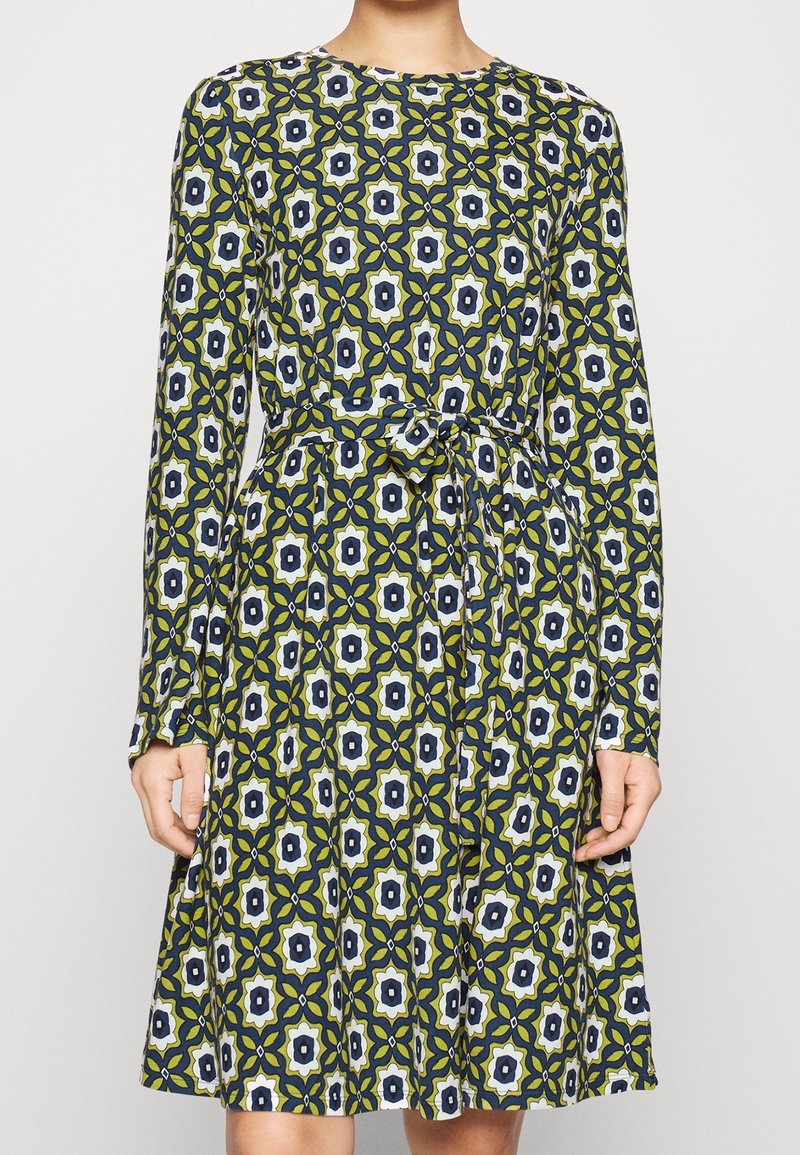 Femme portant une robe à manches longues, longueur genou, avec un motif floral géométrique vert, bleu marine et blanc et une ceinture nouée à la taille.