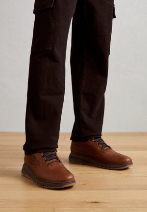 Bruine leren schoenen met zwarte veters gedragen met een zwarte broek, staand op een houten vloer.