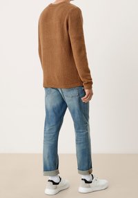 Hnědý texturovaný svetr s dlouhými rukávy, kombinovaný s světle modrými džíny s ohrnutými nohavicemi a bílými teniskami s černými akcenty.