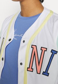 Maillot en maille blanche avec bordure jaune, présentant un "N" rose et un "I" bleu sur le devant. En dessous, un T-shirt bleu avec du texte blanc.