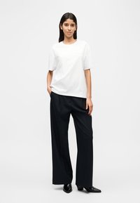 Γυναίκα που στέκεται με τα χέρια στις τσέπες, φορώντας λευκό T-shirt, φαρδιά μαύρη παντελόνα και μαύρα μυτερά παπούτσια, μπροστά σε απλό φόντο.