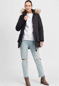 Parka noire avec capuche en fourrure, fermeture éclair et bouton, et deux poches à l'avant. Portée sur un pull blanc et un jean bleu clair déchiré.