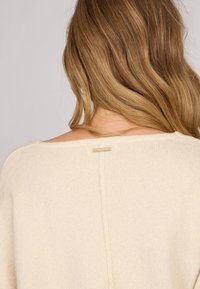 Pull crème au fit ample, avec un col en scoop et une petite étiquette décorative à l'arrière. Tissu tricoté doux et texturé.