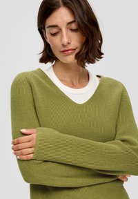 Olijfgroen gebreid sweater met een V-hals, geribbelde textuur en lange mouwen. Gelaagd over een wit T-shirt met een ronde hals.