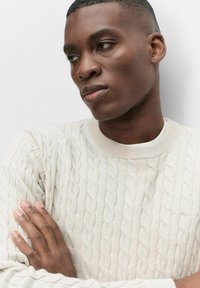 Ljusbeige kabelstickad tröja med ribbad rund halsringning. Slät struktur och subtila sömdetaljer, bärs löst med armarna korsade.