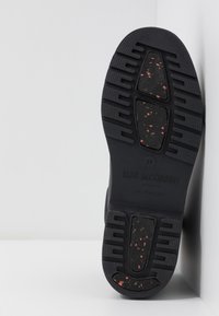 Semelle de chaussure en caoutchouc noir avec une surface texturée et des éclats rouges, dotée de profondes rainures pour une traction optimale et de détails de marque en relief.