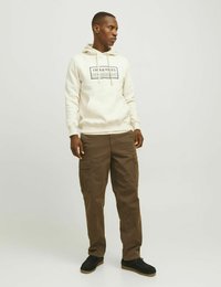 Světle béžová mikina s kapucí a logem; hnědé cargo kalhoty s několika kapsami; černé semišové boty s gumovou podrážkou. Neutrální pozadí.