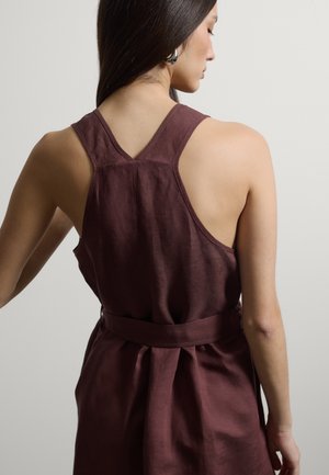 Frau mit langen dunklen Haaren, die ein ärmelloses weinrotes Kleid mit Gürtel trägt, von hinten vor einem schlichten hellen Hintergrund gezeigt.