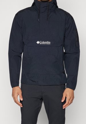 Mand iført sort Columbia Sportswear Company vindjakke med frontlomme og hætte, stående mod en ensfarvet baggrund.