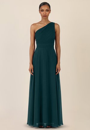 Frau in einem bodenlangen, dunkelgrünen Ein-Schulter-Kleid mit plissiertem Oberteil und fließendem Rock, stehend vor einem einfarbigen beigen Hintergrund.