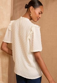 White textured blouse with dotted pattern, ruffled sleeves, and scalloped hem. Worn with dark blue jeans, shown from the back.