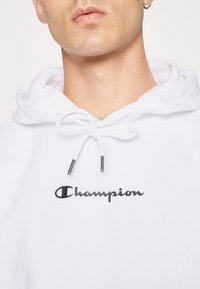 Felpa bianca con cappuccio e coulisse. Presenta un logo "Champion" ricamato in nero sul davanti. Tessuto liscio con vestibilità comoda.
