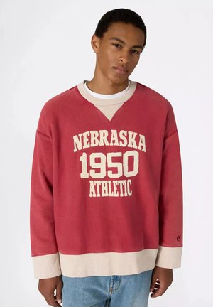 Jonge man draagt een rood-beige trui met tekst "Nebraska 1950 Athletic" en blauwe spijkerbroek tegen een effen witte achtergrond.