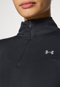 Črna športna zip-up majica iz gladkega, elastičnega materiala. Ima visok ovratnik in majhen srebrn logotip Under Armour na prsih.