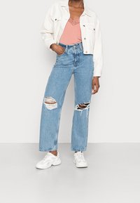 Ljusblå slitna denimjeans, vit kort jacka och en rosa cami-topp med spetskant, kombinerat med vita sneakers.