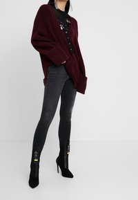 Suéter burdeos de gran tamaño con mangas largas, jeans ajustados negros y botines negros con detalle de cremallera, exhibidos sobre un fondo blanco.