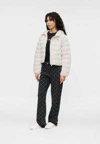 Ελαφρύ κρεμ μπουφάν με φερμουάρ και κουκούλα, φορεμένο πάνω από μαύρο πουκάμισο, συνδυασμένο με σκούρο παντελόνι με σχέδια και λευκά αθλητικά παπούτσια.