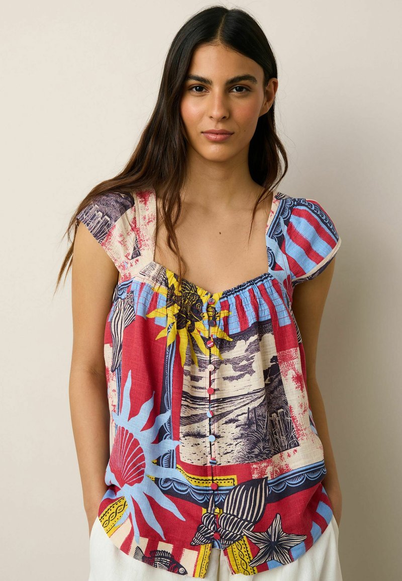 Jeune femme aux longs cheveux noirs portant un chemisier à manches courtes coloré à motifs avec des boutons et un pantalon blanc, debout devant un fond uni.