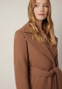 Brązowy, wełniany płaszcz z paskiem, szerokimi klapami i bocznymi kieszeniami. Miękka tekstura z dopasowanym krojem, posiadający fason na zakładkę i prosty krój.