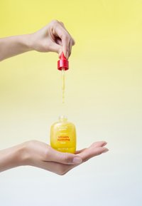 Żółty serum w przezroczystej szklanej butelce z czerwoną zakrętką w formie pipety. Ręka trzyma butelkę, podczas gdy serum kapało z pipety. Miękki gradient w tle.