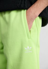 Pantalons de survêtement en coton vert clair avec poches latérales, arborant un logo Adidas blanc et une coupe décontractée. Un pull noir à côtes est visible au-dessus.