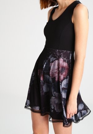 Kobieta ubrana w czarną sukienkę bez rękawów z dopasowanym gorsetem i rozkloszowanym dołem ozdobionym dużymi ciemnoróżowymi i szarymi wzorami kwiatowymi.