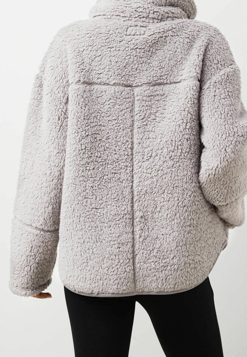 Veste polaire texturée gris clair avec un col montant portée par une personne vue de dos, associée à un pantalon noir.