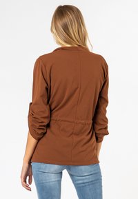 Bruine top met lange mouwen, een relaxte pasvorm en een gathered taille. Gladde stof en opgerolde manchetten, gecombineerd met lichtblauwe jeans.