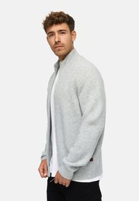 Cárdigan gris acanalado con cuello alto, cremallera frontal y bolsillos laterales, superpuesto a una camiseta blanca. La tela parece suave y texturizada.
