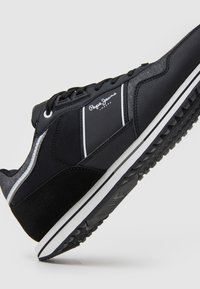 Black athletic shoe with a textured upper, suede accents, and white stripes. Features a rubber sole with a tread pattern for grip.