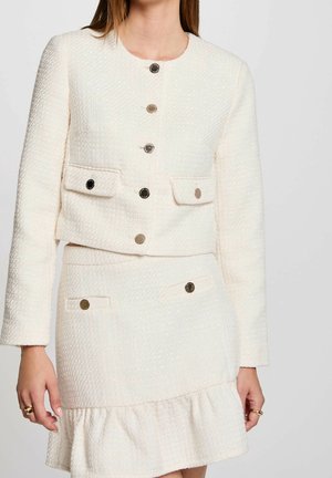 Femme portant une tenue crème en deux pièces texturée avec une veste boutonnée et une jupe assortie à ourlet volanté.