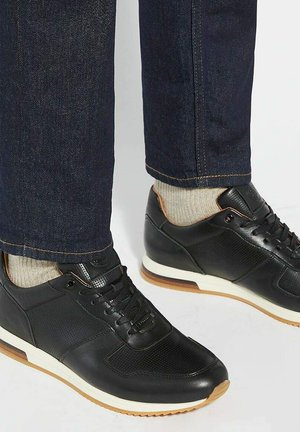 Black athletic shoes with a mix of smooth and perforated leather. White sole, brown accents, and worn with dark jeans and light socks.