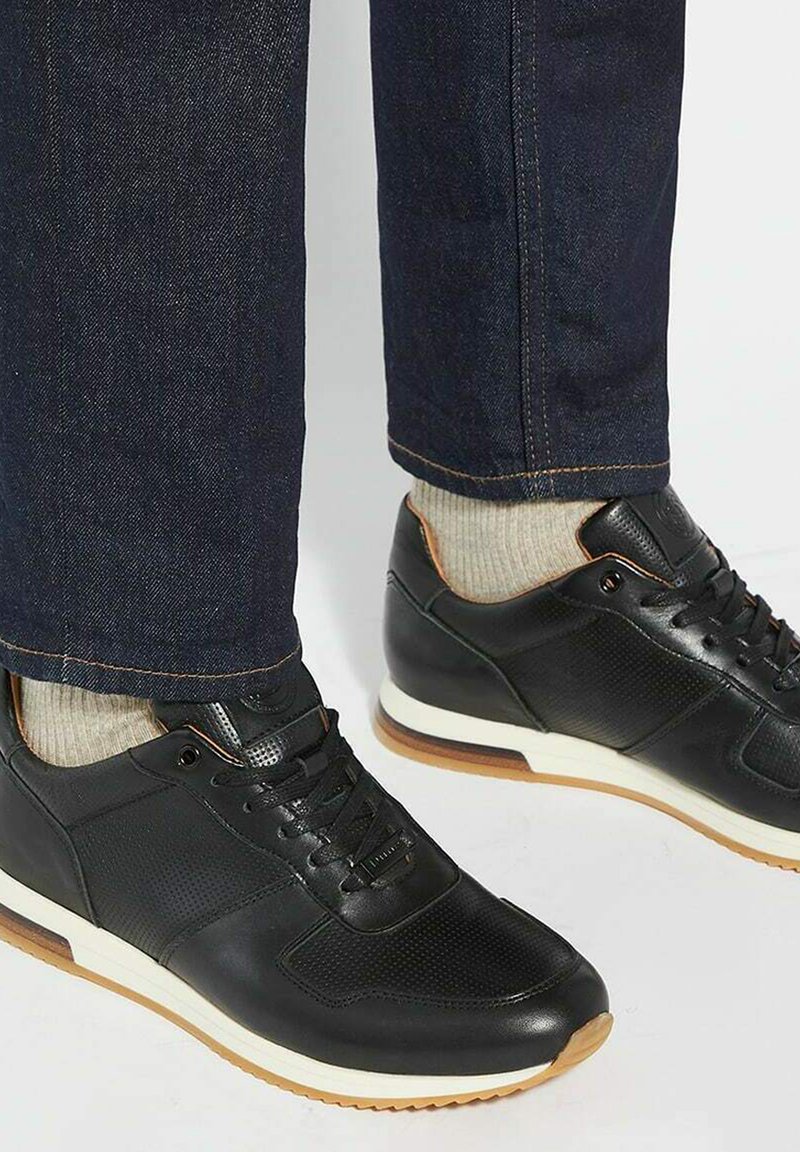 Baskets noires de sport en cuir lisse et perforé. Semelle blanche, accents marron, portées avec un jean foncé et des chaussettes claires.