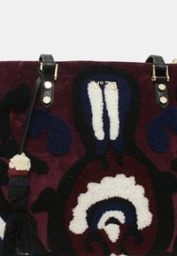 Borsa tote con una superficie burgundy testurizzata, caratterizzata da motivi floreali in blu, bianco e nero. Include manici neri e un pompon decorativo.