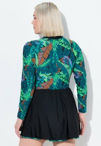Robe à manches longues avec un motif de feuilles tropicales vertes et brunes sur le haut ; une jupe noire s'évase à partir de la taille. Tissu lisse avec une certaine élasticité.
