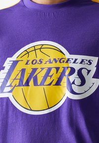 Lila t-shirt med en stor gul basketbollsgrafik och vit text "LOS ANGELES LAKERS". Bomullsmaterial med slät textur.