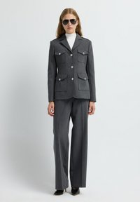 Traje gris ajustado con bolsillos de solapa dobles, cierre de botón y epaulettes. Llevado sobre un suéter de cuello alto blanco y combinado con zapatos de tacón negros.