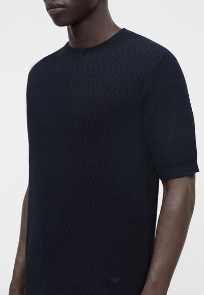 Homme portant un pull en maille texturée bleu marine à manches courtes avec un col rond sur un fond neutre.