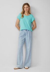 Haut turquoise à manches courtes avec une coupe décontractée, associé à un jeans bleu clair à jambes larges et des baskets beiges. Le haut a un col rond.