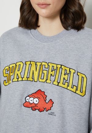 Femme portant un sweat-shirt gris avec "SPRINGFIELD" en grandes lettres jaunes et un poisson cartoon rouge à trois yeux en dessous.