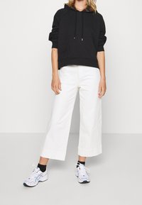 Sudadera negra con capucha y un bolsillo frontal, combinada con pantalones de algodón blancos de pierna ancha y zapatillas blancas con detalles en azul.