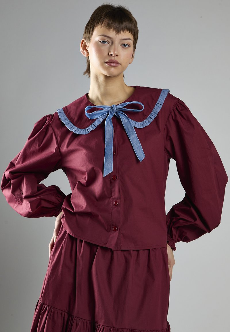 Blusa bordeaux con bottoni, dotata di un ampio colletto a volant, nastro blu a contrasto e maniche a palloncino. Tessuto liscio con una leggera lucentezza.