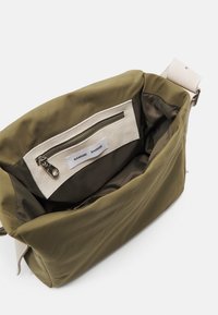Bolso de hombro de tela verde oliva con una textura suave, bolsillo interior con cremallera y acentos color crema. Forma rectangular con correa ajustable.