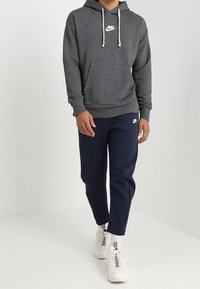 Grå huvtröja med vit Nike-logga, kombinerad med marinblå joggingbyxor. Vita sneakers med mönstrade snören fulländar outfiten.