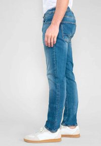 Un jean en denim bleu clair avec une coupe slim, comportant cinq poches et un léger délavage. Associé à des baskets blanches et une chemise blanche unie.