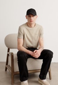 Μπεζ βαμβακερό μπλουζάκι, μαύρα παντελόνια και μαύρο καπέλο φορεμένα από ένα καθιστό άτομο σε μια ανοιχτόχρωμη, στρογγυλή πολυθρόνα με ξύλινα πόδια.